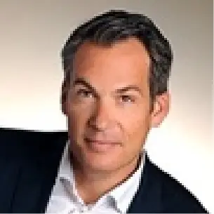 Man with dark hair wearing a white shirt collar and dark suit jacket against a light background