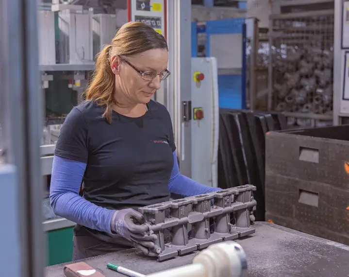 Frau mit langem Haar und Handschuhen hält ein graues Metallteil in einer Werkstatt mit Maschinen im Hintergrund