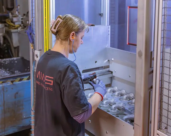 Person mit zusammengebundenem Haar und Handschuhen bedient eine Maschine mit mehreren Metallteilen in einer Werkstatt