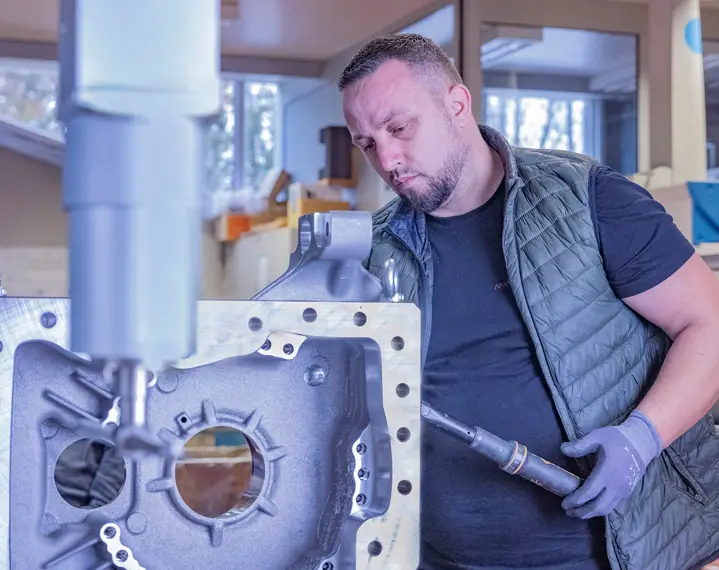 Mann mit Weste und Handschuhen bedient eine Maschine an einem großen Metallteil in einer Werkstatt