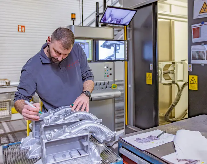 Mann mit grauem Pullover arbeitet an einem metallischen Maschinenteil in einer Werkstatt mit Monitoren und Warnschildern im Hintergrund